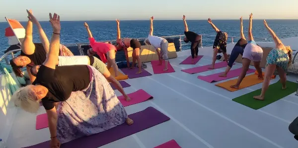 Skupina lidí na palubě lodě v jóga pozicích a stretching cvičeních při západu slunce.