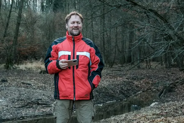 Dan v červené bunda stojí v zimním lese s mobilem a usmívá se.